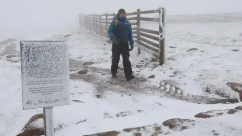 Peter Jolly/Northpix A mountain ranger wearing a winter jacket, hat and boots walks through the snow. There is a snow-covered sign and a wooden fence