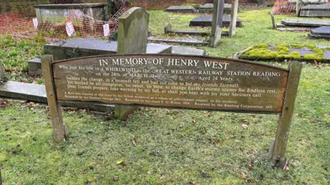 Wooden memorial which reads "In memory of Henry West, who lost his life in a whirlwind at the Great Western Railway Station Reading on the 24th March 1840. Aged 24 years.
“Sudden the change, I in a moment fell and had not time to bid my friends farewell.
“Yet hushed be all complaint, ’tis sweet, ’tis blest, to change Earth’s stormy scenes for endless rest.
“Dear friends prepare, take warning by my fall, so shall you hear with joy your Saviour’s call.” The memorial is in front of gravestones in a graveyard