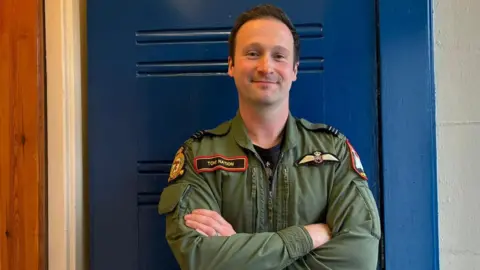 A man with short brown hair pushed back has his hands folded and is smiling into the camera and standing against a blue door. He is wearing a khaki green uniform which says 'Tom Nation' on one side with a logo on the other.