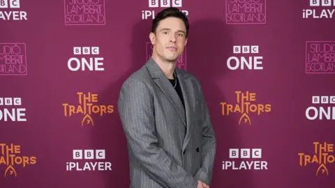 PA Media Ed Gamble wearing a grey pinstriped jacket and black T-shirt looks at the camera with his hands folded 