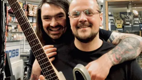 Sam Orr is smiling as he stands behind Mark Wood in Sam's guitar shop. Mark Wood is also smiling as he holds his guitar, with the neck of the guitar and the frets visible.