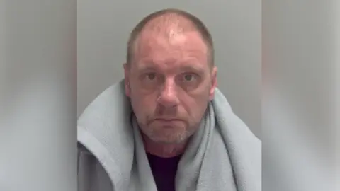 Elvis Vickers, a shaven headed man stares at the camera in a police mugshot. He has a light grey fleecy blanket over his shoulders and is wearing a dark blue T-shirt.