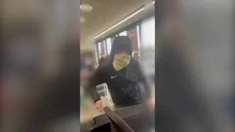 Leo Carpin wearing a hooded black Nike jacket and a scarf over his mouth, holding a knife in his hand and leaning over a supermarket checkout.