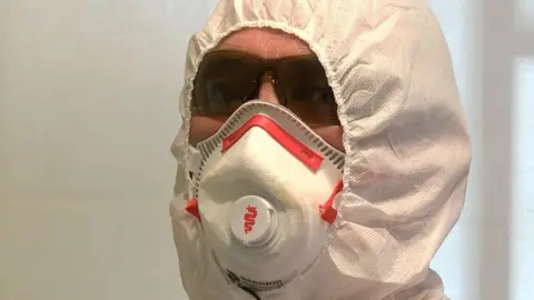 The photo is a close up of trauma cleaner, Jim Gildea's face. There is little skin visible as he's wearing a forensic suit, face mask and protective glasses.