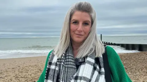 Nicola is standing on a beach and wears a dark green woolly long sleeved top and a scarf with large black and white checks. She has shoulder length straight greying hair, pulled back from her face at the front.