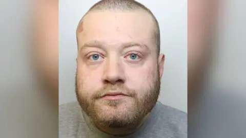 Avon and Somerset Police Christopher looks at the camera with a blank expression. He has light hair, shaven very short, and a light beard, and wears a grey prison jumper. 