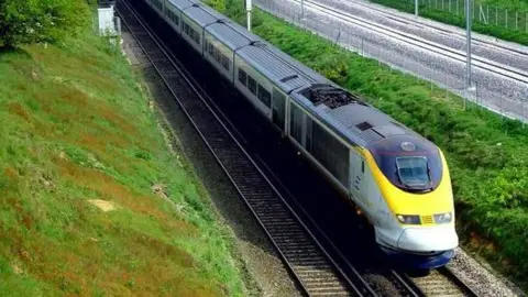 PA Media A train with a yellow front, on tracks surrounded by banks of green grass