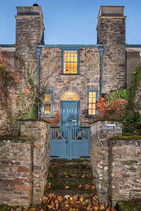 Historic England Archive The entrance to Sharlands House, an Edwardian townhouse. There are granite steps leading up to two blue wooden gates and then leading up to a blue wooden door. There are narrow windows either side of the door and a larger window above it. Lights are on inside. There are two large granite chimneys. 