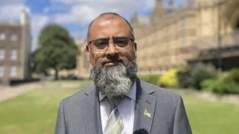 Iqbal Mohamed looking serious while wearing a grey suit, with a small pin badge that has a Palestinian flag on it and stood in a grassy area buildings in the background