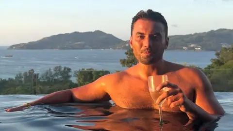 Jordan Wright holding a wine glass at the edge of an infinity pool. He has brown hair and stubble and the sun is shining on him. Tall green hills and the sea are pictured behind him.