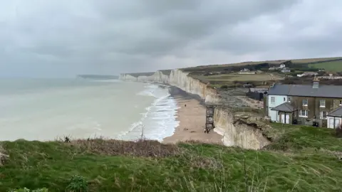 A beach on an overcast day. Some houses are on top of its white cliffs. 