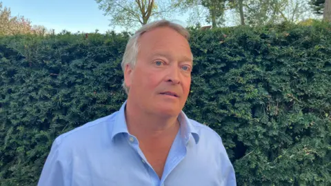 Vikki Irwin/BBC Nick Green is wearing a light blue, collared shirt and is standing in front of a dark green hedge. You can see blue sky and trees in the background. 