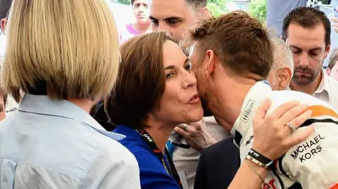 Getty Images A woman with short dark brown hair in a crowd, kissing a man wearing racing gear on the cheek