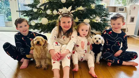 Family photograph Left to right: Joshua, dog Bonnie, Chloe, Sophie, dog Bailey and Jack all sitting by a Christmas tree with large white baubles. The children are all smiling. There is a wooden toy kitchen to the left and French doors behind them leading to the garden. 