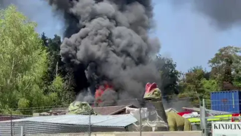 Black smoke and orange flames are seen coming from the unit. There are some model dinosaurs in the foreground behind a barbed wire fence. 