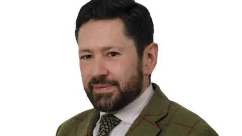 A man in his 40s with a beard and dark hair. He is wearing a tweed jacket, shirt and tie.