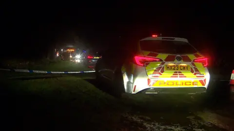 A police car on the grass at the side of the road next to a police cordon.