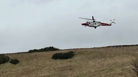 Fife Jammer Locations A red and white helicopter hovers in a grey sky above a field. Four bushes below and yellow-brown grass.