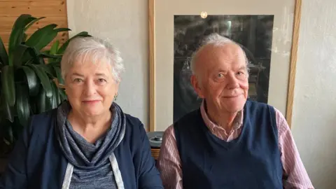 A woman and a man with grey hair looking at a camera in front of a plant and picture in a frame
