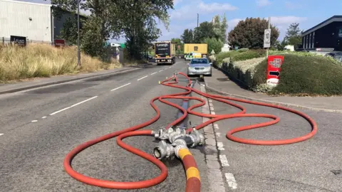 Firefighter equipment on the road next to the scene of a large fire. The blaze cannot be seen in the shot, but there is a large hose pipe and a police van blocking off access to the road.