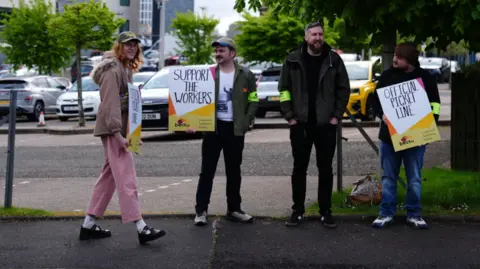 PA Media Four people stand on a pavement near an office building holding picket signs reading “Support the Workers” and “Official Picket Line”. Some wear fluorescent armbands as they gather beside trees and a car park on an overcast day.