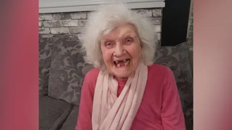 An elderly woman is sat on a grey sofa. She is smiling widely and has white hair with lots of volume and is wearing a pink jumper and pale pink scarf.