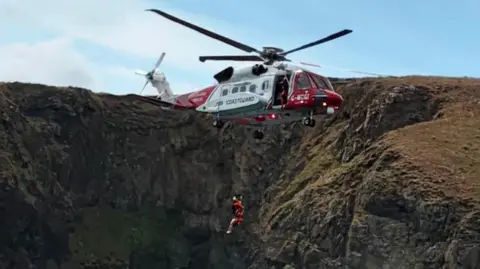The coastguard helicopter is winching two people very close to a rocky cliff face.
