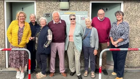 BBC A group of eight volunteers stand outside a pebble dash toilet block. They are mostly elderly and a mix of men and women. On the toilet block wall is a large Welsh flag. The toilet block has two doors, one for men and one for women, and a grey slate roof, with two railings in front of it. The woman far left has a leopard print dress in the colours of pink and cream, and white trainers, over her dress is a yellow puffer coat, she has gold-framed glasses and short blonde hair. Next to her is a woman with blonde hair tied up and a blue coat and trousers. Next to her is a woman with a plaid shirt with a blue gilet over it, and blue jeans. She has cream Birkenstocks on and short grey hair. Next to her is na man with a dark pink jumper over a pink shirt, and pink trousers with brown shoes, he has white hair, and a receding hairline. Next to him is a woman with shades on and short grey hair, she is wearing a peach top with a light-green cardigan, and cream trousers. Next to her is a woman with a blue striped t-shirt and grey cardigan, she also has blue trousers on and the brown strap of a handbag across her shoulder. Next to her is a man with a pink jumper and brown trousers, he is bald. And on the right is a woman with her arms in front of her, she has a flowery dark blue top on with dark blue jeans. She has short grey hair.