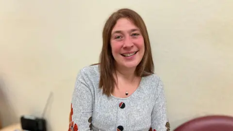 A woman is smiling at the camera. She has long brown hair and a grey top.
