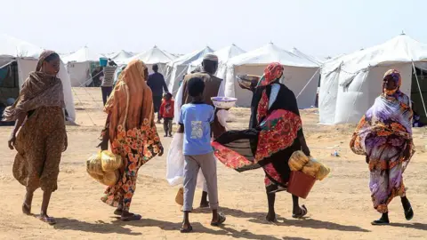 AFP via Getty Images Four women, a boy and a man walking on sandy ground carrying food. In the background are white aid tents.