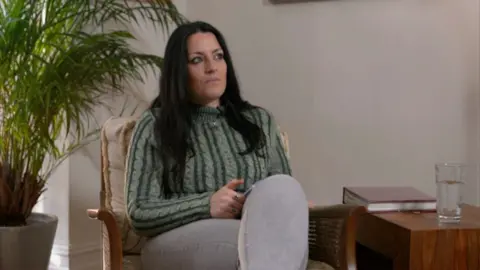 Rebecca has long dark hair and is wearing a green and cream polo neck sweater. She is sitting on the stairs with her hands on her lap. There is a plant with big green leaves behind her. She is not smiling.