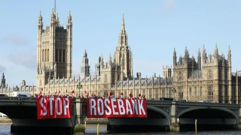 Getty Images A large red sign with white letters spelling out STOP ROSEBANK being hung of the side of a bridge in front of Westminster