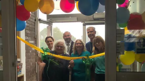 BBC A balloon arch behind automatic doors. There is a yellow ribbon with green bows. Six people are stood behind the ribbon. The woman on the front left is holding scissors ready to cut the ribbon.
