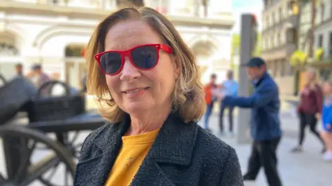 Clodagh Keating has red glasses, a yellow top and a navy coat. She's got blond hair, worn short and smiles at the camera