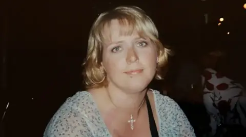 A picture of Clare Wood, who is wearing a light blue top, a crucifix around her neck and has blonde hair. She is looking at the camera.