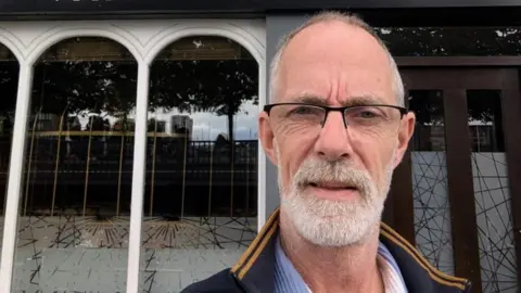 Mal has a long face, well groomed grey beard and wearing thin rectangular glasses. The collar of his sports jacket is turned up. 