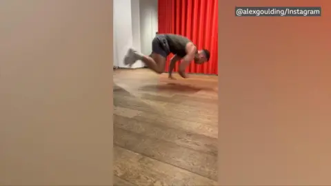 Alex Goulding is mid-air jumping off the floor after doing a push-up in a plank position. He is wearing a dark green tank top, shorts and white trainers. He is performing the push up in front of a red curtain. 