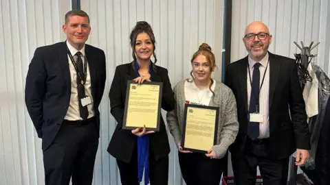 South Yorkshire Police Two women smile and hold up framed certificates. They are flanked on either side by men in suits.