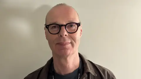 Emma Craig/BBC Jan Pitman standing against a light coloured wall. He is wearing round dark-framed glasses and a brown shirt over a black T-shirt. 