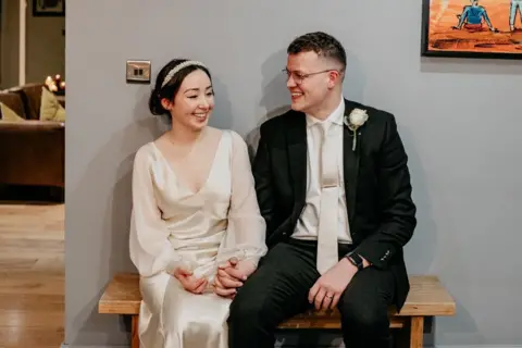 Ben and Olivia are sat on a bench at a wedding, Olivia in a white wedding dress hand in hand with Ben who wears a suit with a matching coloured tie. They are both smiling and looking at each other.
