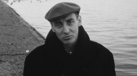 Spike Milligan standing by a pond wearing a black coat and looking directly into the camera.