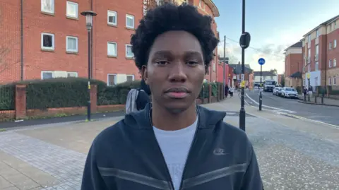 Michael is wearing a grey zip-up hoodie. He is standing on a paved walkway in a residential-style area with red‑brick buildings behind.