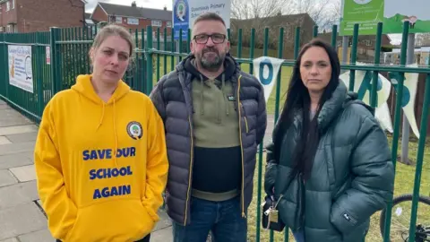 BBC/Seb Cheer Two women and a man stand by a green fence outside a school looking at the camera. One of the women is wearing a yellow hoodie with the words 'save our school again' printed on the front. A sign in the background reads 'Queensway Primary School'.