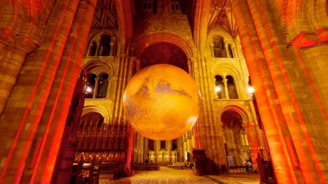 Photograph of the inside of Peel Catherdral. A large Mars planet sculpture is hanging from the ceiling, while the walls and floors are lit a bright orange.