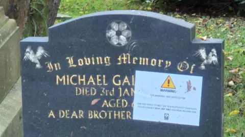 BBC A  black gravestone with gold lettering at Riddrie Park Cemetery - the first name of the deceased is Michael, but the surname has been covered by a large sticker saying WARNING with a exclamation mark in a yellow box   