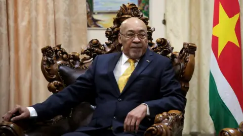 Reuters President Desi Bouterse sitting in a wooden chair with a red flag beside him