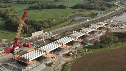 HS2 Viaduct in countryside, showing seven spans each made up of four light grey beams supported on pillars. A large red and yellow crane is visible in the foreground.