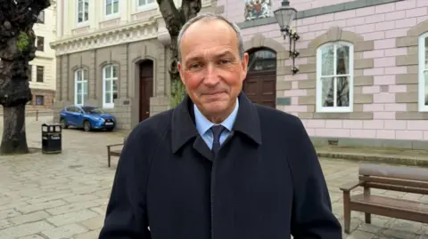 Deputy Lyndon Farnham is wearing a black coat, blue shirt and blue tie. He is pictured in front of the States Assembly building. 