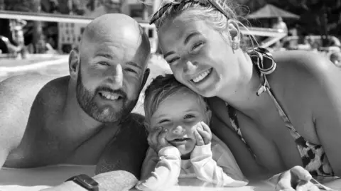 Warren Davies A black and white photo of a man and a woman with a youn girl between them. They are in a row, leaning up on the side of a swimming pool. The man is bald, with a beard, and is shirtless. The woman has ligth hair piled on her head with sunglasses perched on top. She is wearing a swimsuit. The little girls is resting her chin in her hands with her elbows propped up on the side. They are all smiling and looking at the camera.