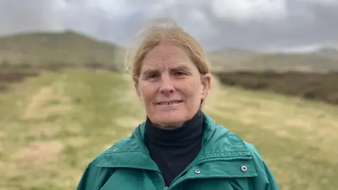 Tracy May is wearing a green waterproof coat, standing on Dartmoor. There is a long strip of cut moorland - that acts as a firebreak - in the background. Either side of this is gorse, heather and other vegetation. The sky is grey and cloudy.